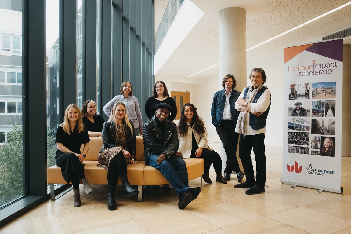 A group of nine people sitting on a sofa at a university open space. They are next to a project poster with our acknowledgement logo on it