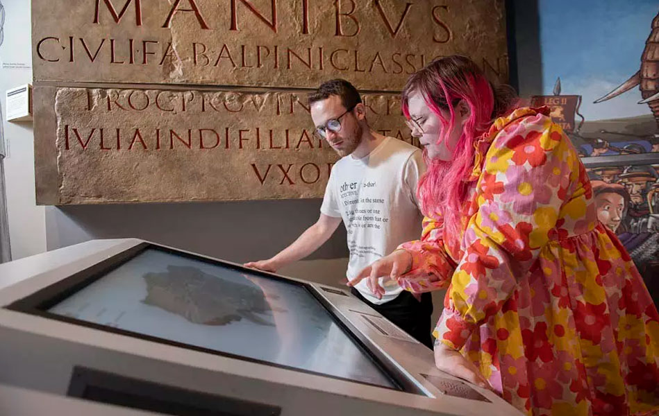 Two people using a digital display screen in a museum, with a large carved stone behind them