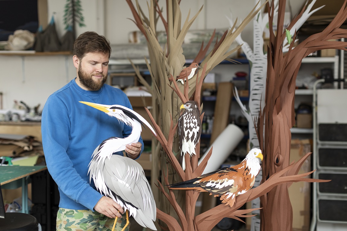 A person standing with life-like paper sculptures of birds and trees, such as a grey heron