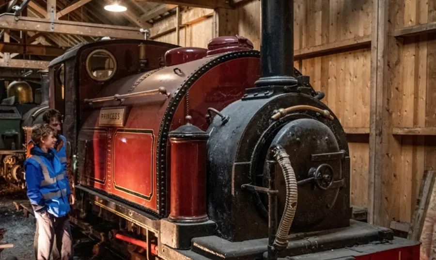 A behind-the-scenes tour at the Ffestiniog and Welsh Highland Railway. Two visitors investigate a red tank engine in a wooden shed at the Ffestiniog and Welsh Highland Railway.