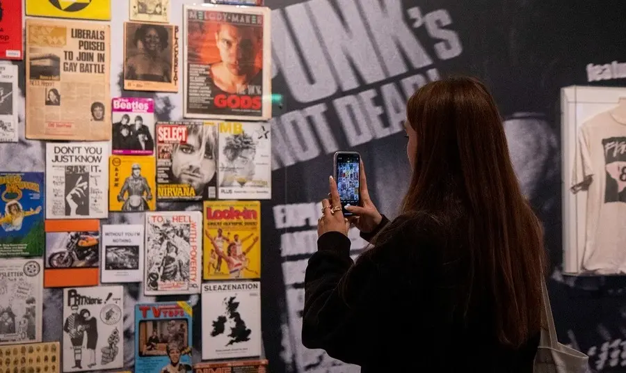 Display of gig posters at The Museum of Youth Culture Image of person with long dark hair and dark clothing taking a picture on a mobile of a display of music gig posters at The Museum of Youth Culture