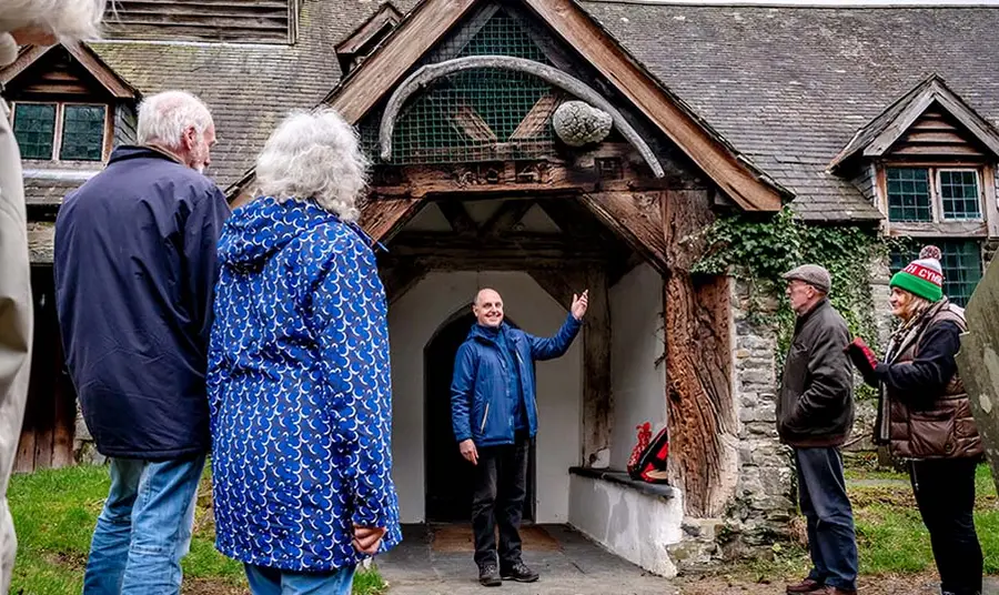 Croesawu ymwelwyr i Eglwys Sant Tydecho yng Ngwynedd, Cymru. Credyd: Ymddiriedolaeth Eglwysi Genedlaethol Golygfa awyr agored o berson yn croesawu grŵp o bobl i eglwys