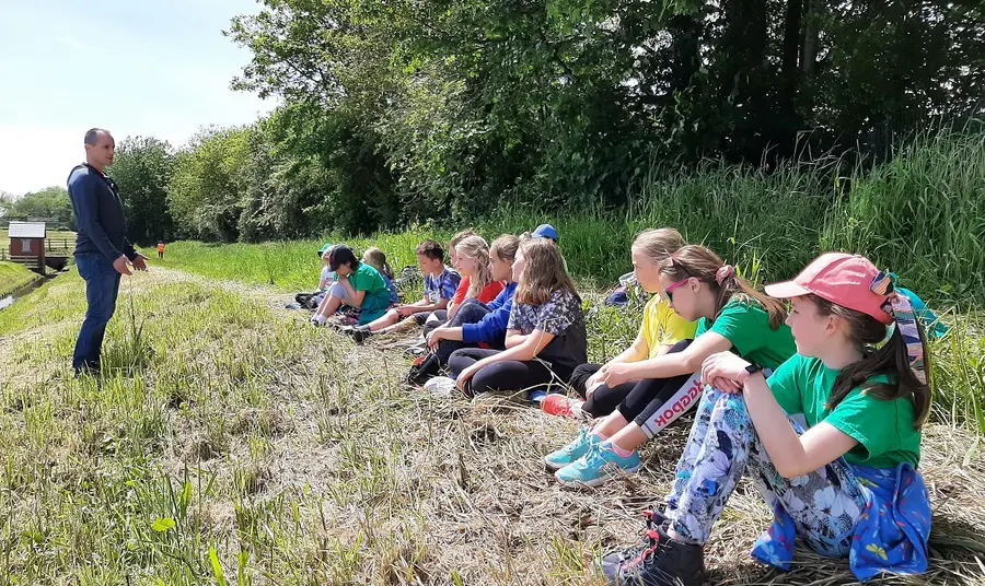 A group of children sitting down in front of a row or trees, listening to someone giving a talk