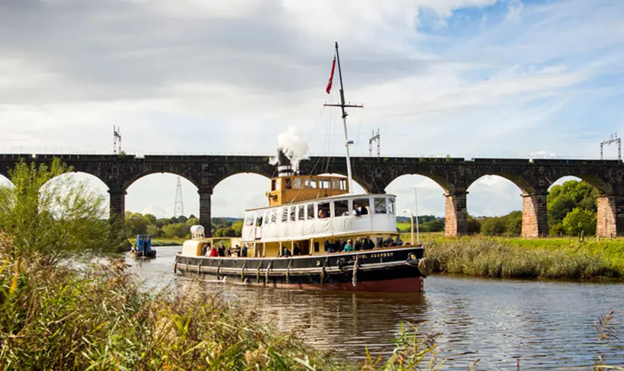 A boat on a waterway with a bridge in the background and plants visible on the banks of the water