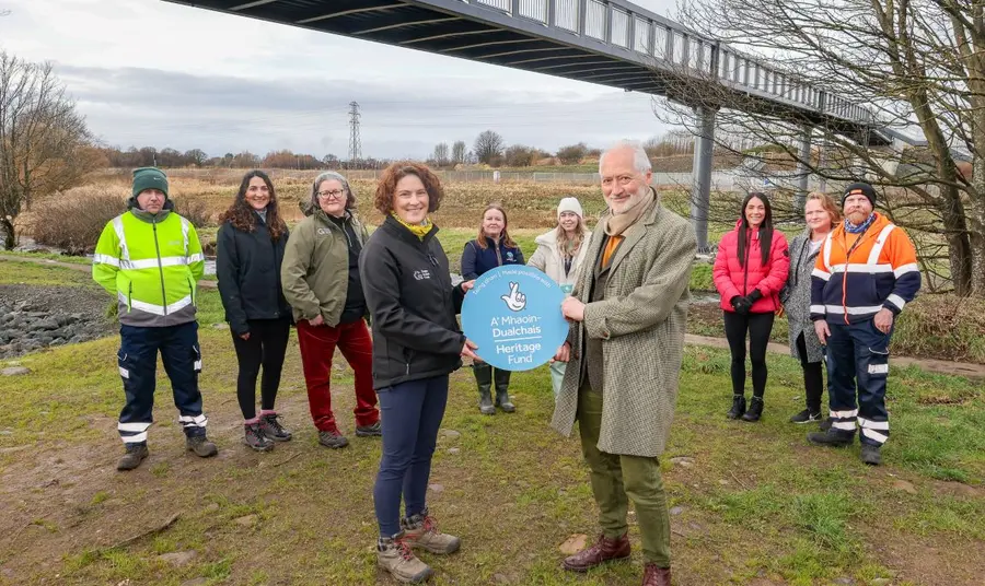 A group of organisations, local and national, are working together on the project. A group of representatives from different organisations involved in the River Park project gather together on the banks of the river, with a pedestrian bridge crossing behind them. To the front, two hold a Heritage Fund roundel.