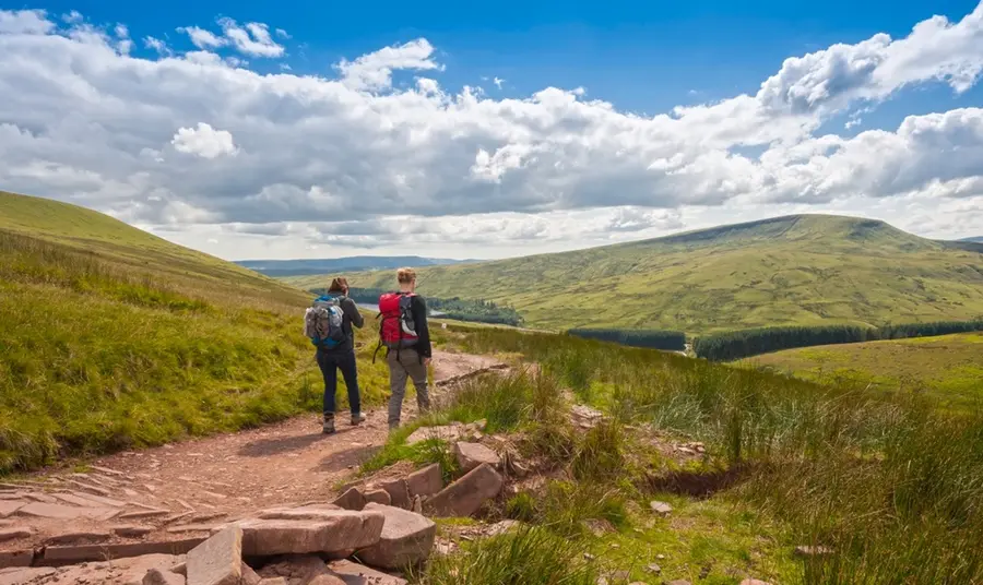 Hikers in the Brecon Beacons National Park. Two hikers walk away down a dirt track into an open, green expanse of the Brecon Beacons, on a sunny day.