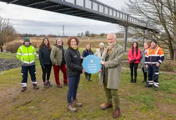 A group of representatives from different organisations involved in the River Park project gather together on the banks of the river, with a pedestrian bridge crossing behind them. To the front, two hold a Heritage Fund roundel.