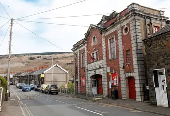 The red brick facade of Tylorstown Welfare Hall. The building rises above surrounding terraced houses. The hills of the Rhondda can be seen over the rooftops.