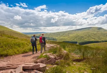 Two hikers walk away down a dirt track into an open, green expanse of the Brecon Beacons, on a sunny day.