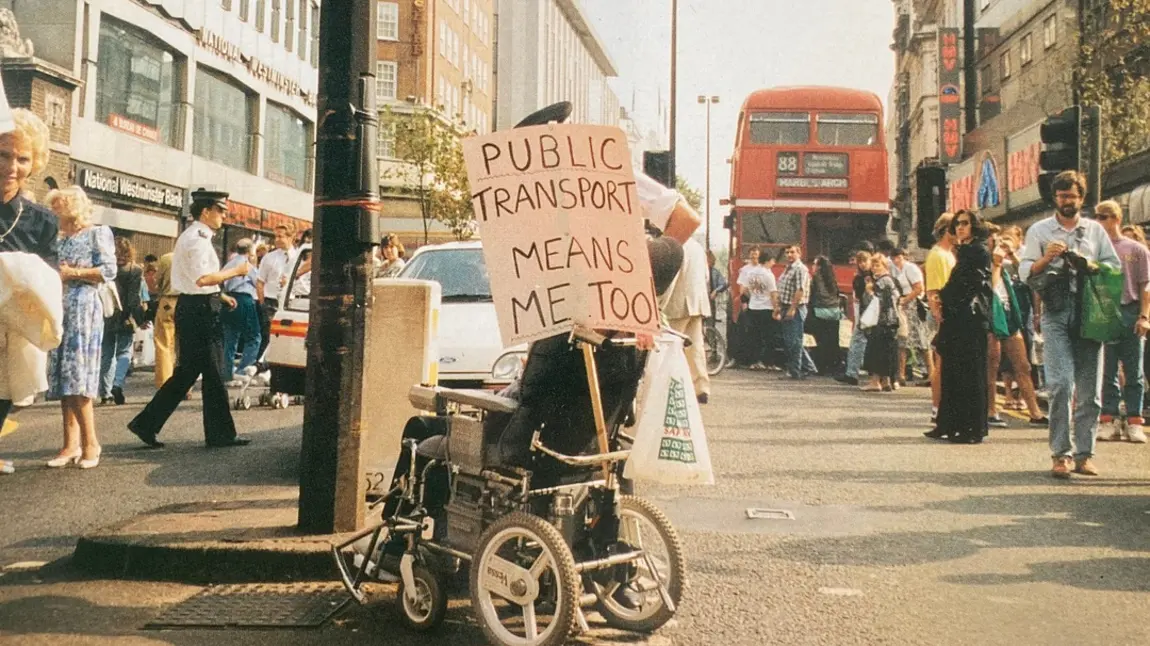 Image of disabled person in 1990s, protesting with plaque saying public transport means me too Image of disabled person in 1990s, protesting with plaque saying public transport means me too