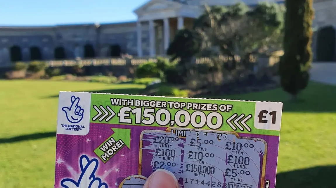 A National Lottery scratchcard held in front of a large building with classical architecture and lawn