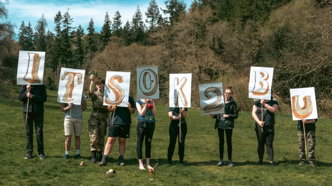 Image of people standing in a line in a countryside setting holding signs reading It's OK 2 B U