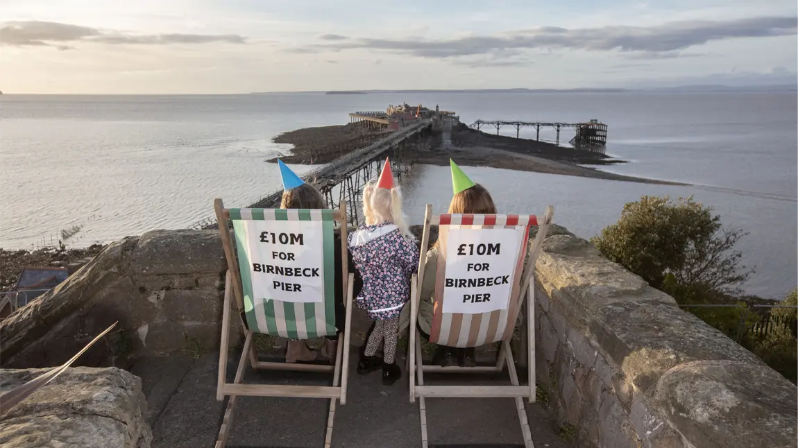 People sitting on deck chairs wearing party hats in front of Birnbeck Pier