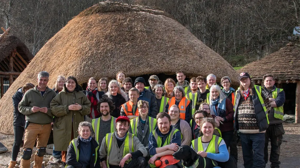 A group of people some wearing high-vis jackets stand together in front of a round house made from wood with a thatch roof