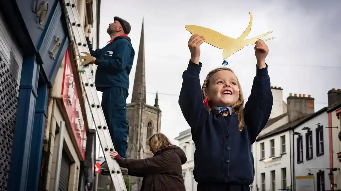 A young girl holds a cut out of a golden bird above her head. In the background are two adults working on a ladder.